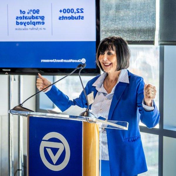 A person standing a podium smiles while gesturing with their hands. The screen behind them contains statistics saying 22,000+ students and 90% of graduates employed or in further education.
