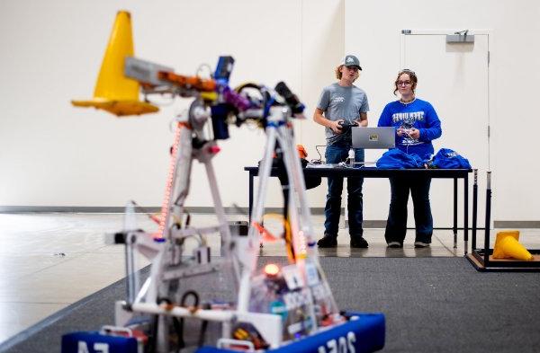 two people stand at table, robot from FIRST Robotics in the foreground